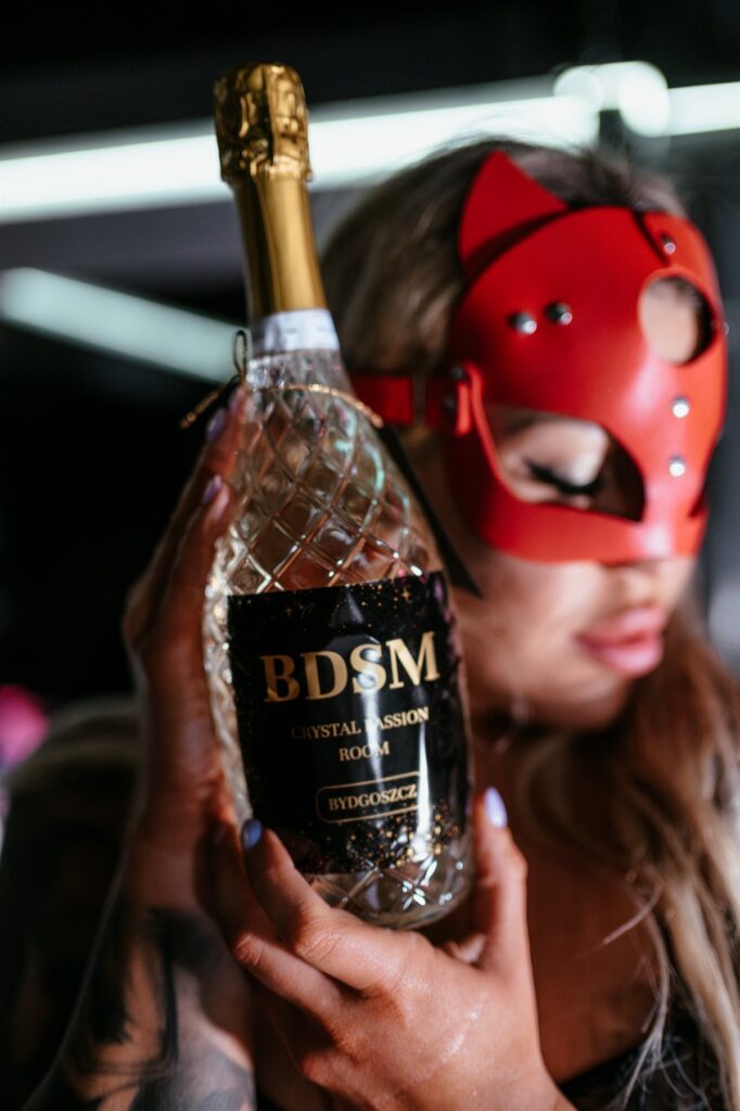 Kobieta w czerwonej masce trzyma butelkę alkoholu z napisem BDSM
